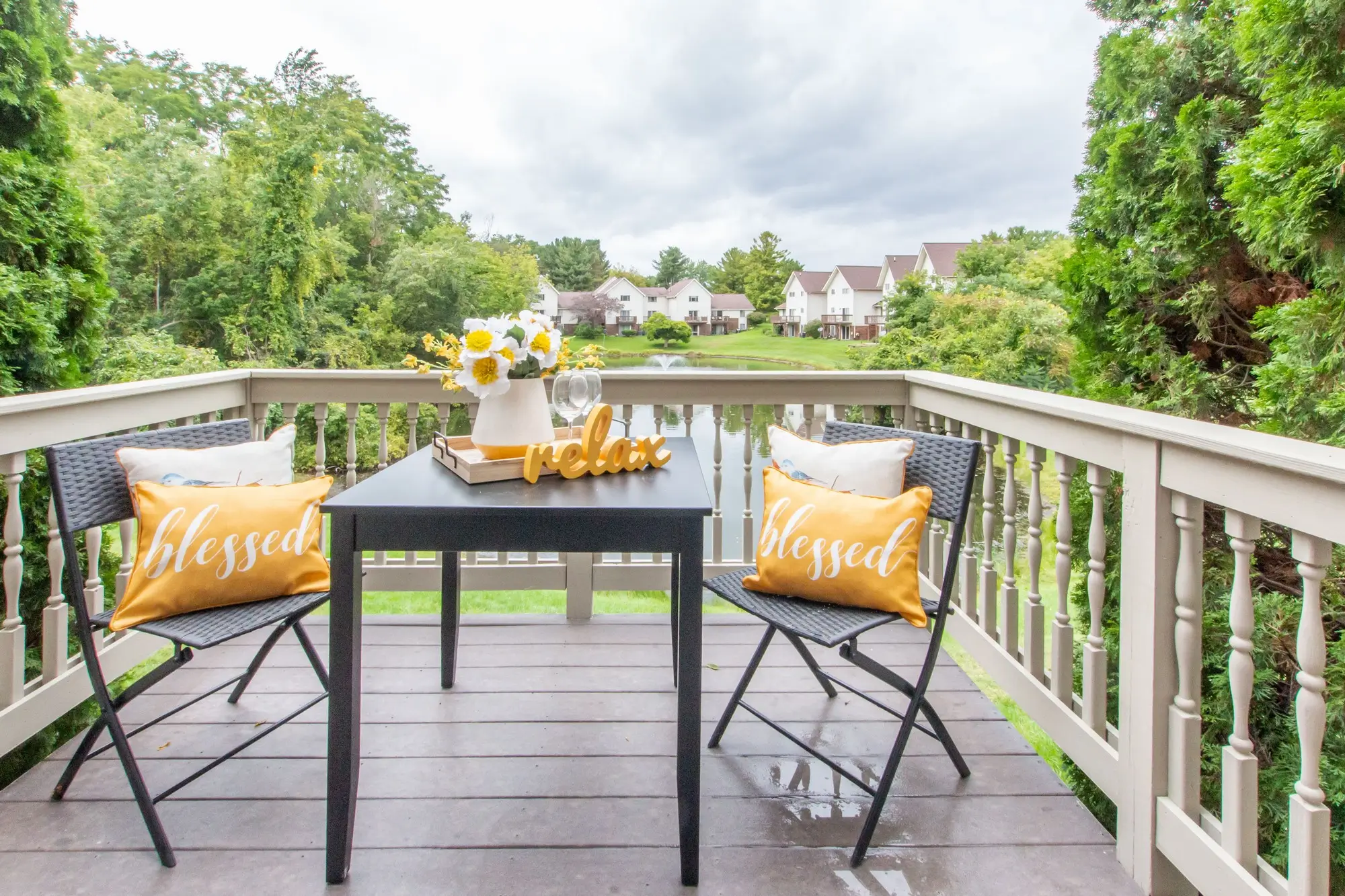 Private balcony of a rental townhome at Bavarian Village on the Lake in Clarkston, MI, featuring an outdoor patio table set overlooking a scenic community pond and fountain.